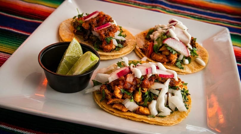 Three vibrant chicken tacos with radishes, onions, and sauce, served with lime wedges on a white plate.
