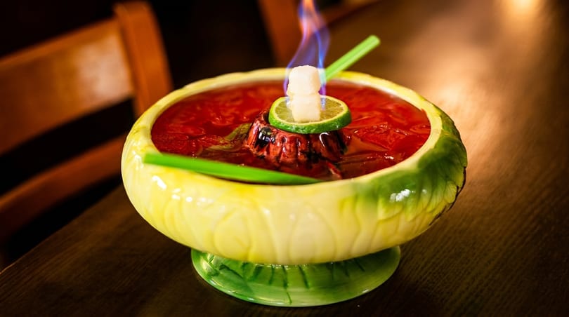 Flaming red cocktail in a green and yellow bowl with a lime and sugar cube garnish.