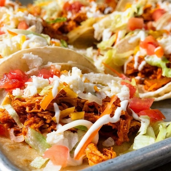A tray of chicken tacos topped with lettuce, tomatoes, cheese, and a drizzle of white sauce.