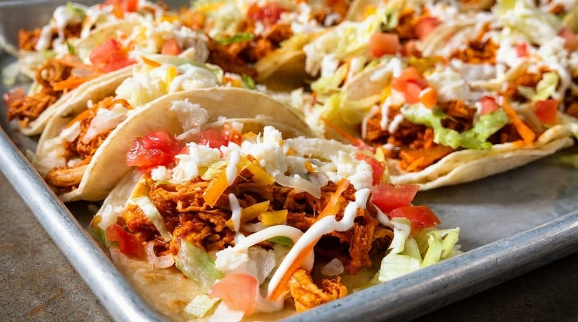 A tray of chicken tacos topped with lettuce, tomatoes, cheese, and a drizzle of white sauce.