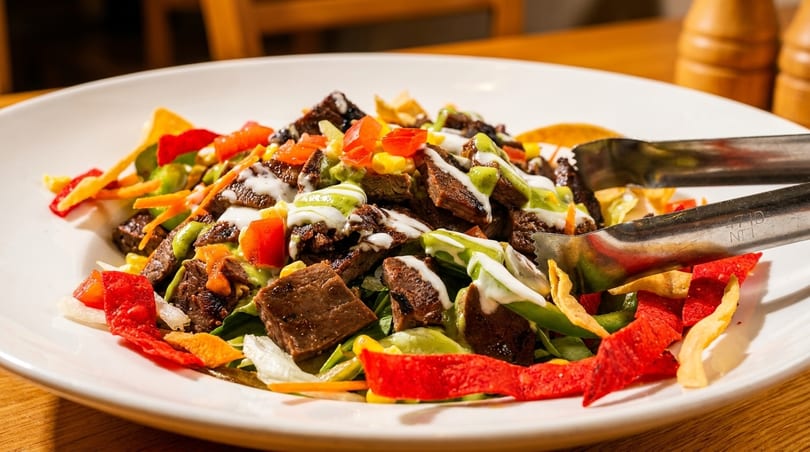 Steak salad with corn, tomatoes, colorful tortilla strips, and green and white drizzles.